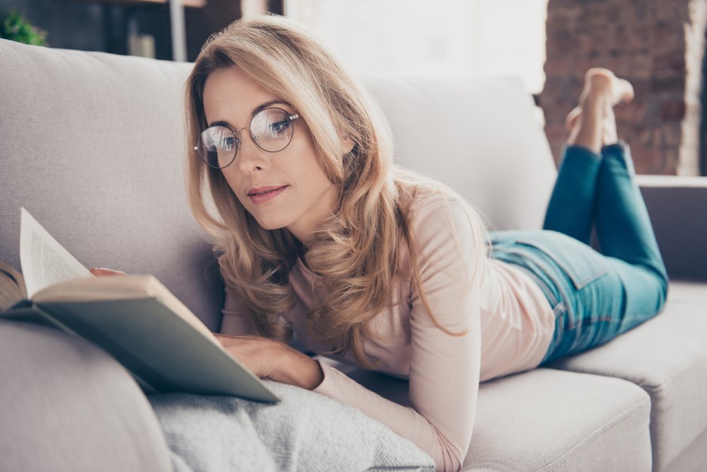 young lady laying down on her couch reading a book with her feet up