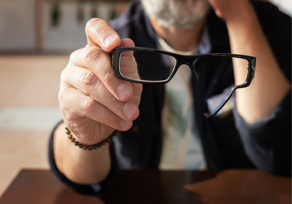 An elderly man with a gray beard shows broken glasses, holding his head with his hand