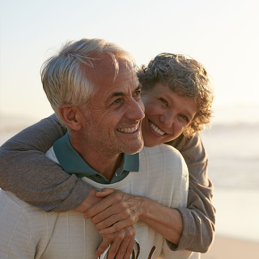 Portrait-of-happy-mature-man-being-embraced-by-his-wife-at-the-beach.-Senior-couple-having-fun-at-the-sea-shore.