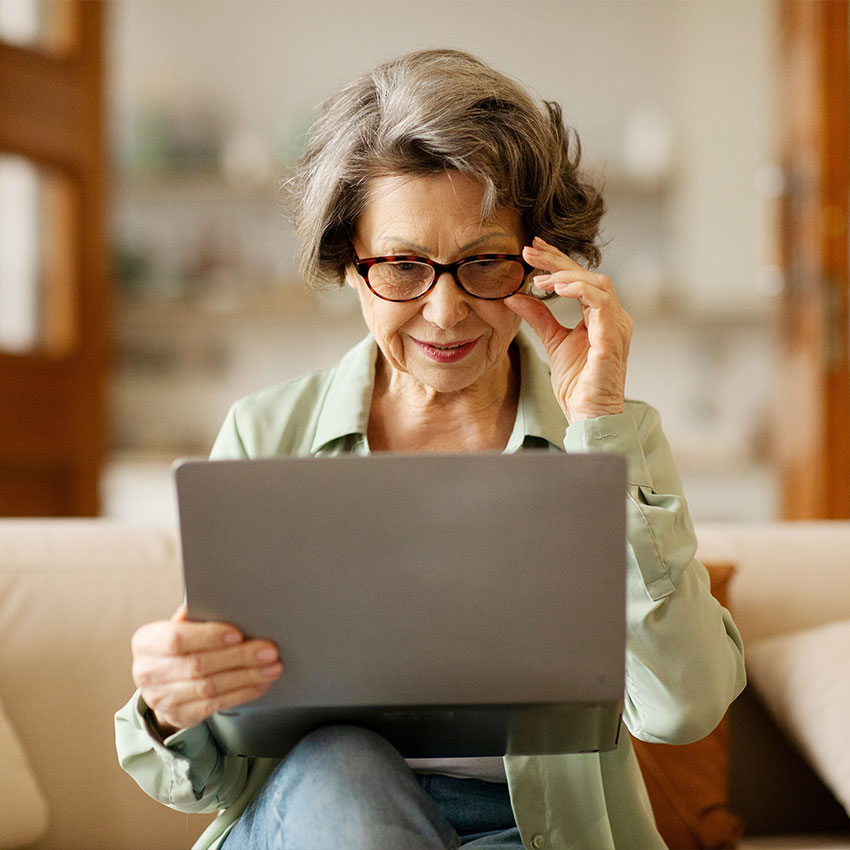 Positive-senior-woman-using-laptop-and-touching-glasses,-surfing-internet-or-video-calling-with-relatives,-sitting-on-sofa-at-home-interior