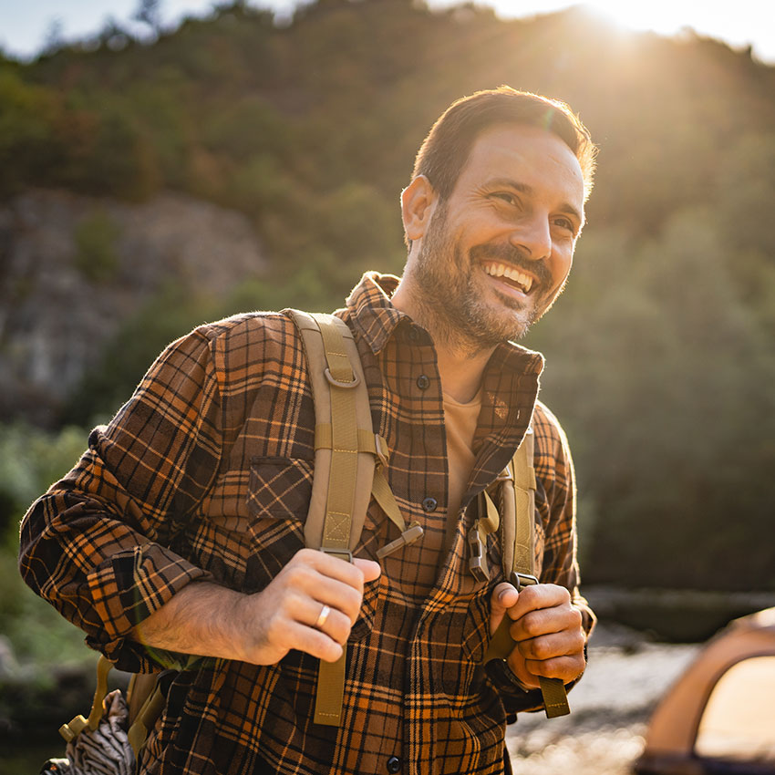 adult-man-hiker-wearing-a-large-backpack-on-a-camping-trip,-standing-near-a-tent-and-a-folding-chair-by-a-river,-surrounded-by-lush-forest-and-hills-during-sunrise