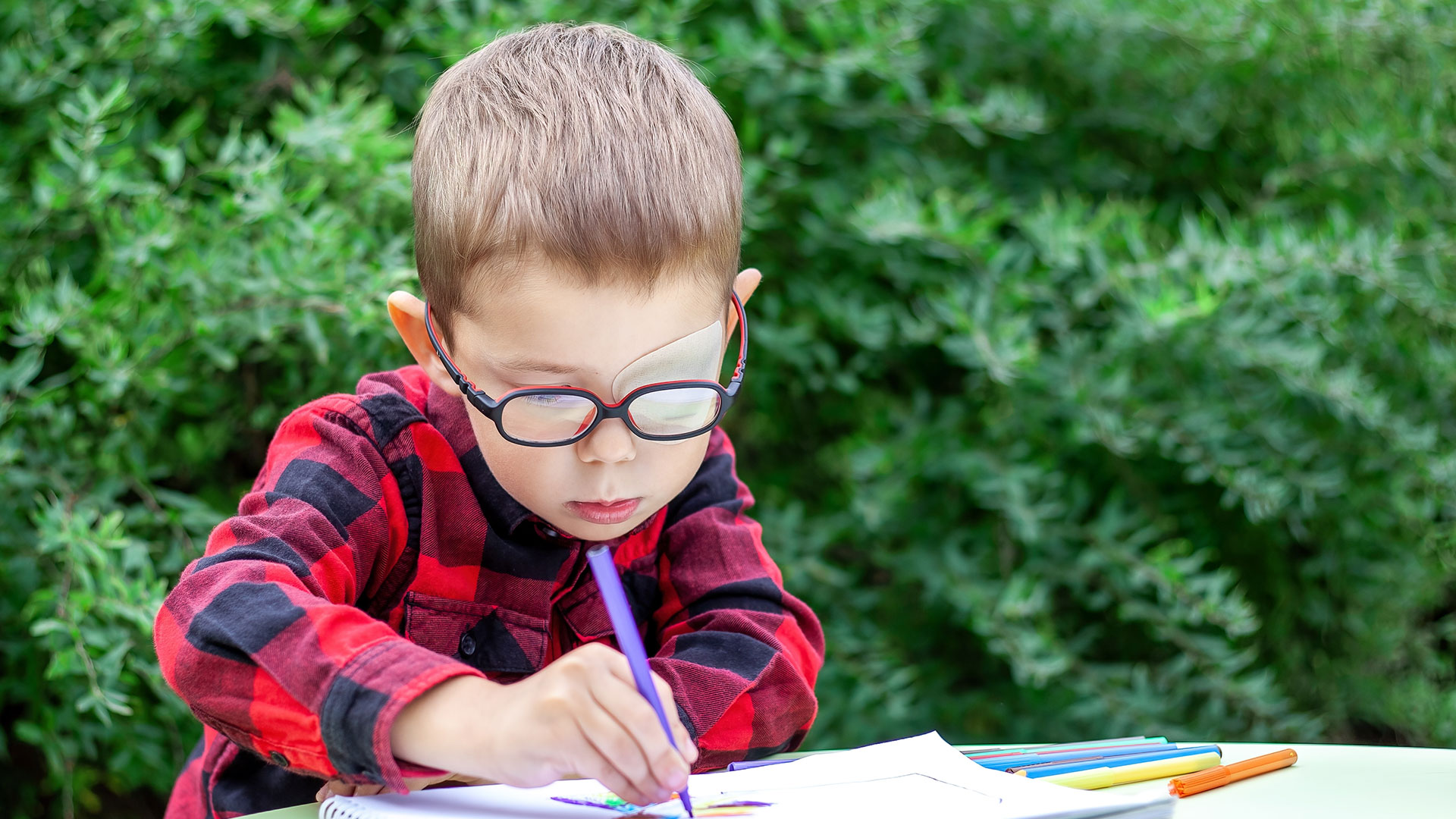 A-little-boy-draws-in-the-garden-(outdoors).-He-wears-glasses-and-an-eye-patch-(occluder)-to-prevent-amblyopia-and-strabismus