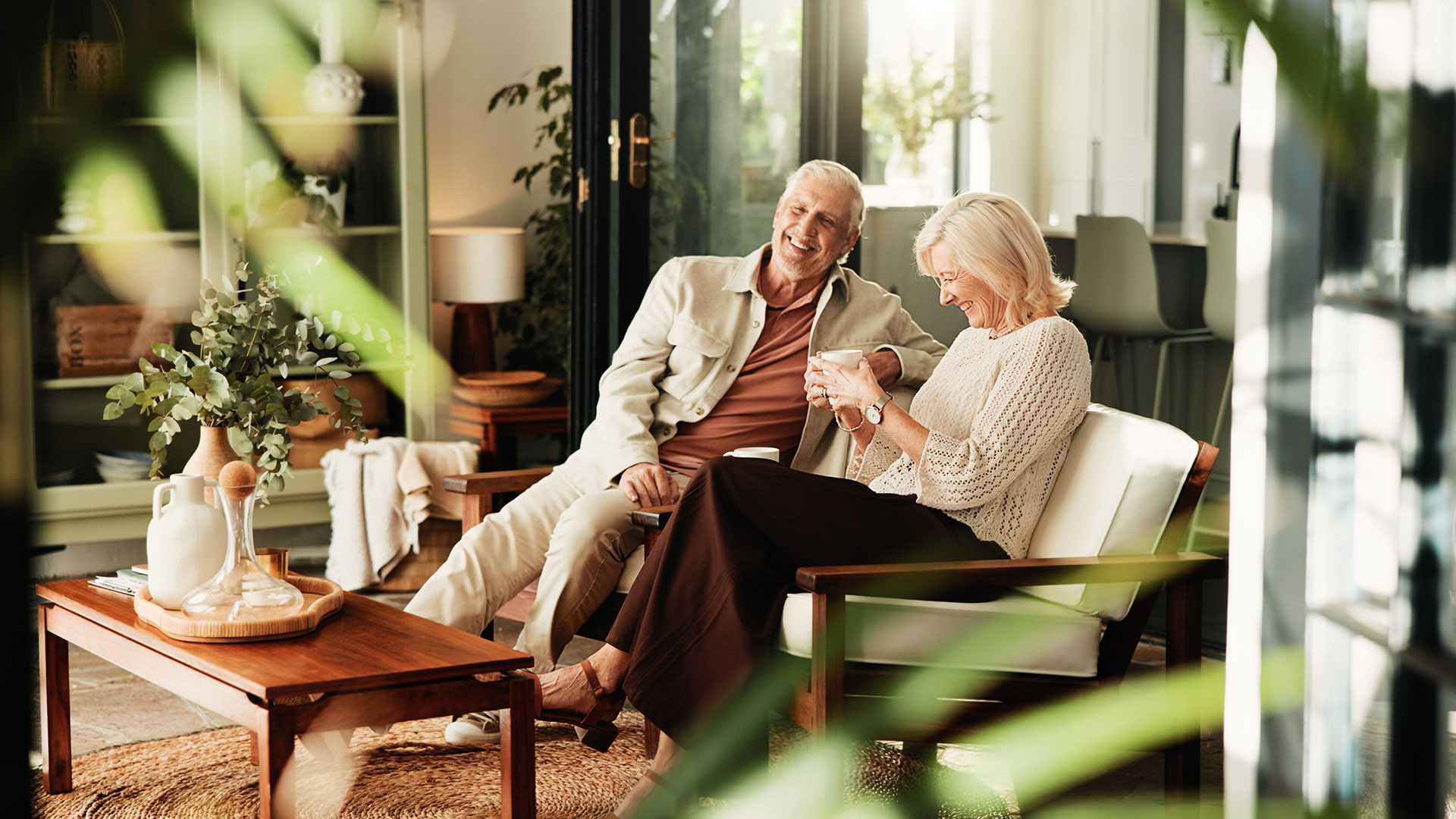 Coffee,-laughing-and-relax-with-old-couple-in-living-room-of-home-together-for-break-or-wellness