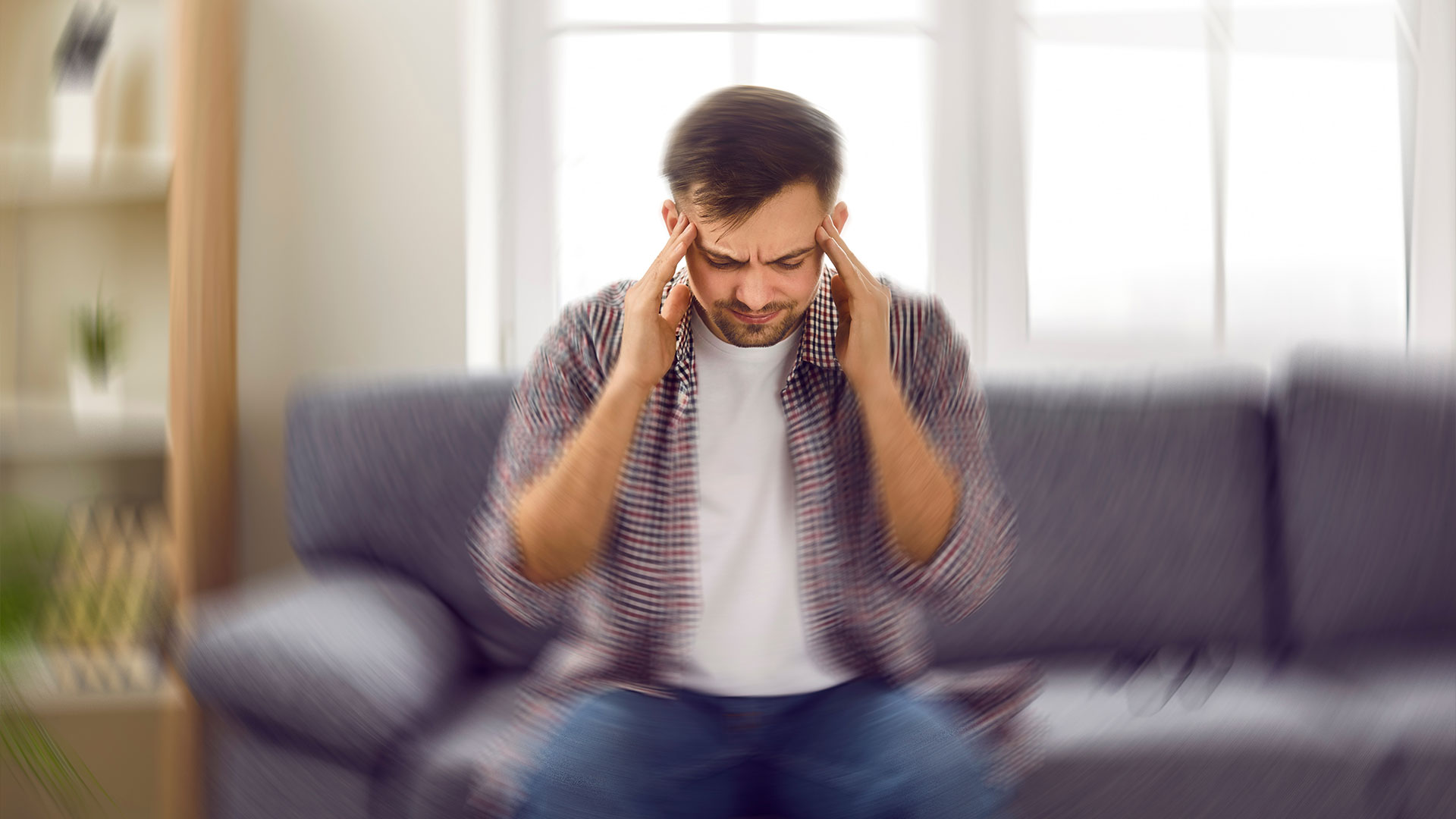 Man-suddenly-feels-dizzy-and-takes-a-seat-on-the-sofa.-Young-guy-feeling-pain-and-spinning-sensation-in-his-head