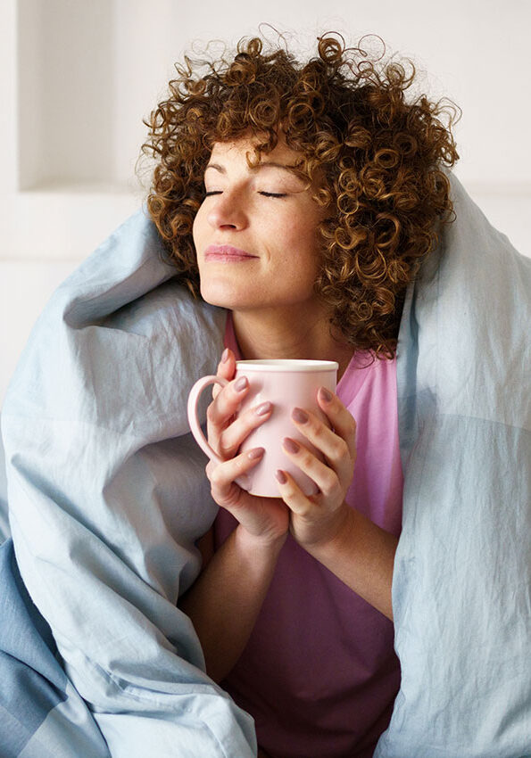 Relaxed-curly-woman-with-closed-eyes-wrapped-in-blanket-while-holding-mug-of-hot-beverage-on-bed-in-weekend