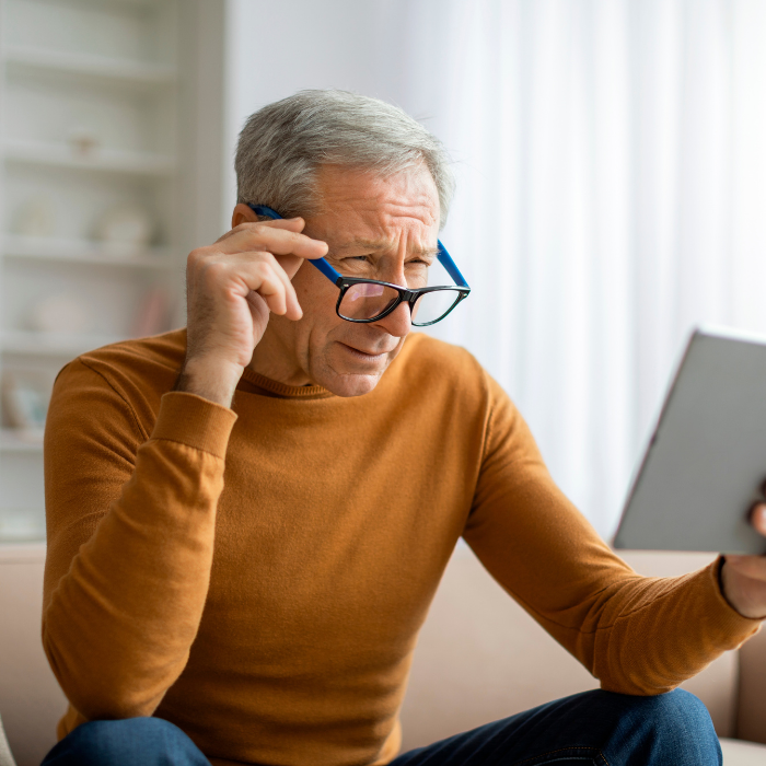 Senior retired grey-haired man grandfather adjusting his eyeglasses while using tablet at home, sitting on couch, reading news
