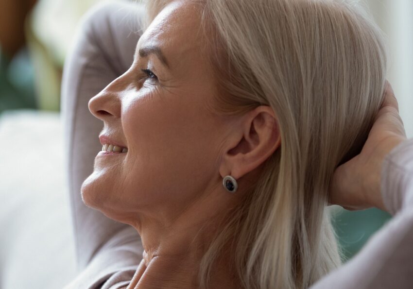 Elderly woman sitting on couch with hands behind head smiling and looking up
